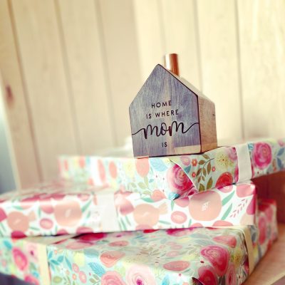 A little wooden house with a copper chimney sits on a pile of presents engraved with the words "Home is where Mom is."