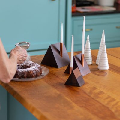 A hand sprinkling powdered sugar on a cake with three a-frame candlestick holders and three little wooden trees sitting behind the cake on a counter.