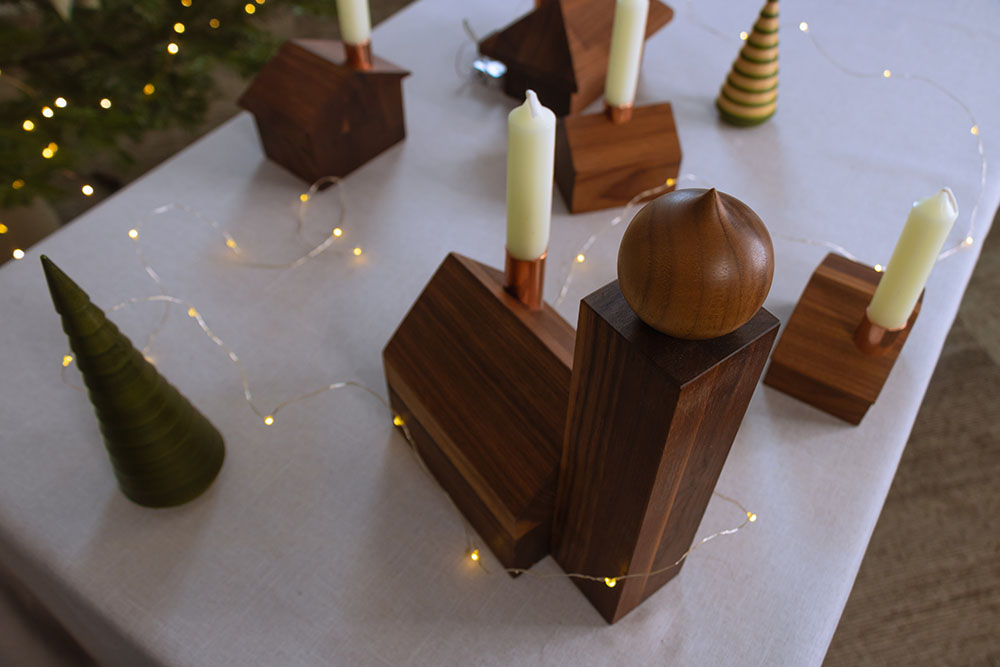 A handcrafted Alpine Church ornament with a hand-turned onion dome and copper chimney is displayed on a linen-covered table. Surrounded by wooden house candle holders, fairy lights, and decorative trees, it creates a cozy, festive atmosphere.