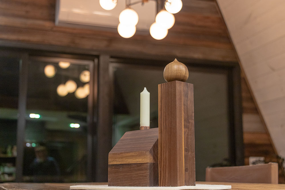 A wooden Alpine Church ornament with an onion dome and a copper chimney holding an unlit candle sits on a table. The warm, wood-paneled interior and softly glowing lights in the background enhance the cozy atmosphere.