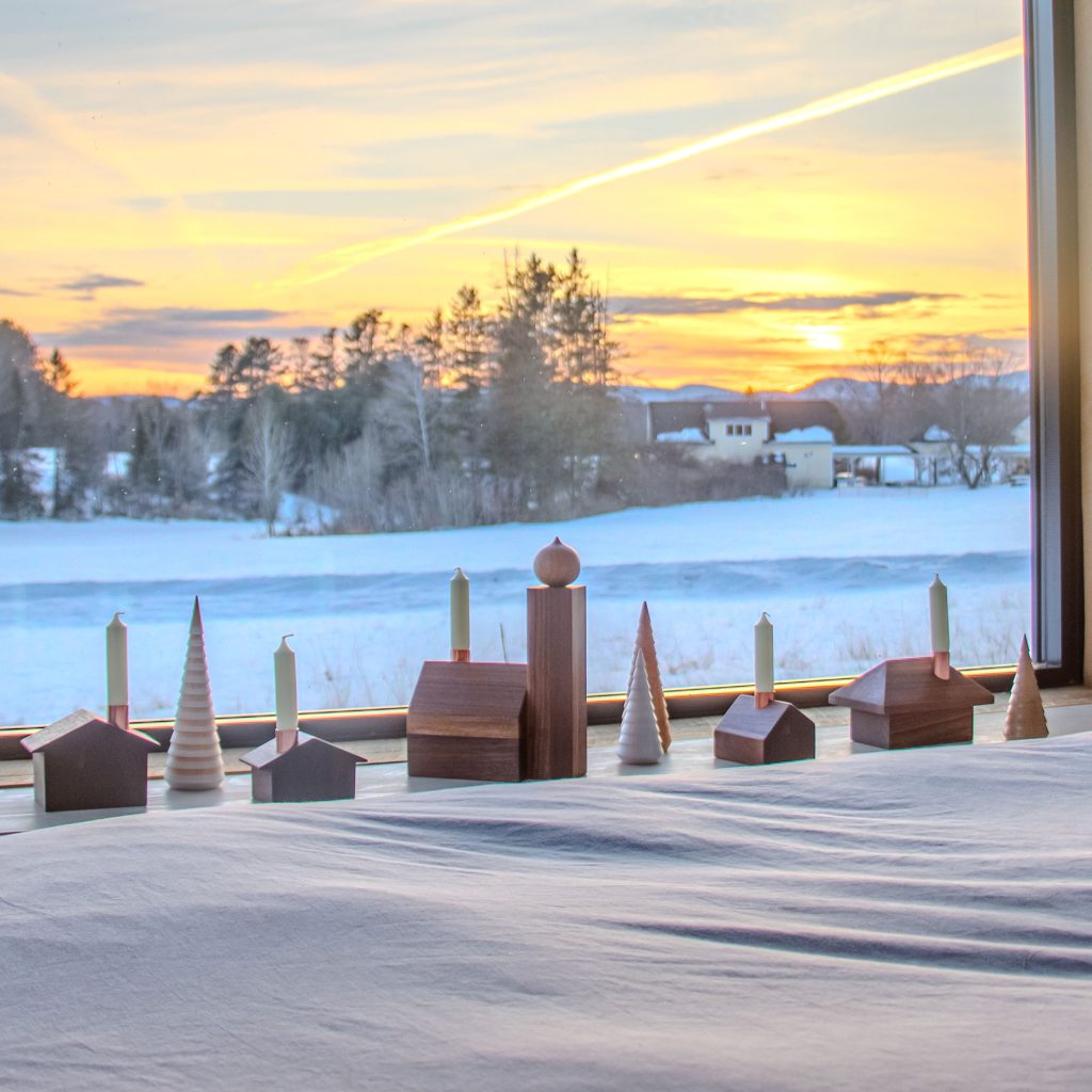 A wooden alpine village display sits on a windowsill in a cozy bedroom, overlooking a snowy landscape at sunset.
