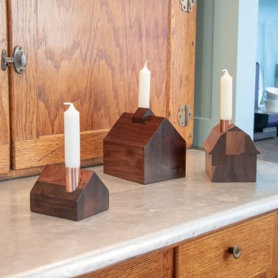A set of three tiny wooden house candlestick holders on a countertop with white tapered candles sticking out of their copper chimneys.