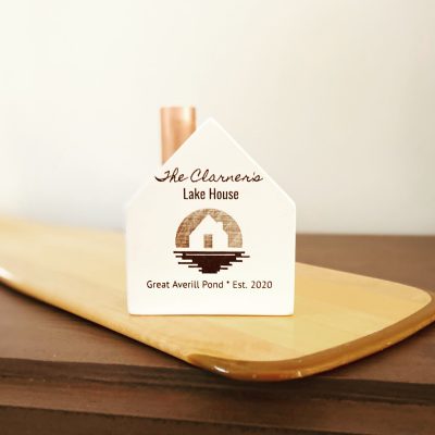 A little white wooden house candlestick holder with a copper chimney and a custom engraved decorative image and the words "Lake House, Great Averill Pond, Est 2020."