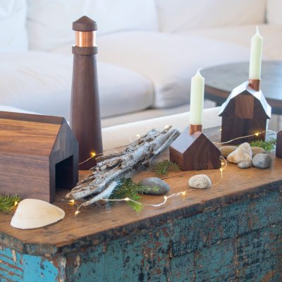 A decorative modern coastal collection of little wooden structures on a shelf with some rocks, driftwood, shells, and fairy lights.