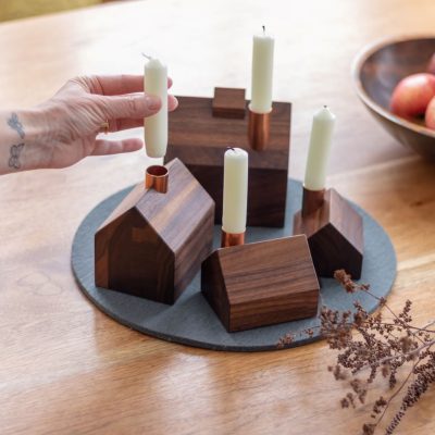 Four little wooden house candlestick holders with white tapered candles in their copper chimneys sit on a gray felt pad next to a bowl of fruit and decorative twigs.