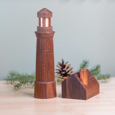A wooden lighthouse with a copper lens and a Saltbox house candlestick holder with a copper chimney, set against a light background with pine accents.