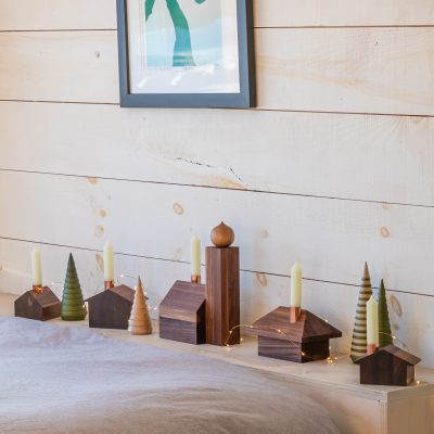 A wooden alpine village display sits on a light wooden surface against a shiplap wall. Copper chimneys hold unlit candles, while fairy lights weave through the arrangement, creating a cozy, festive ambiance.