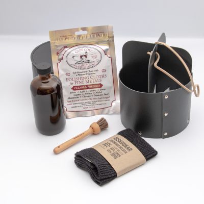 Bottle of tung oil, a wooden cleaning brush, a black organic linen rag and foil bag of two metal polish clothes sit next to a black cardboard storage cylinder with lid