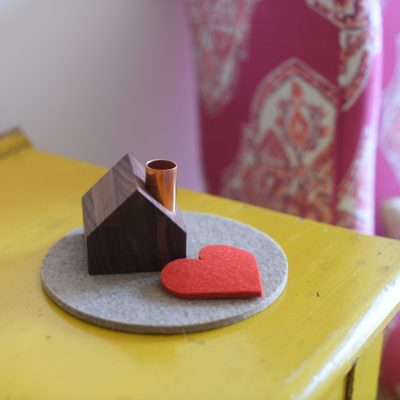 A tiny wooden house with a copper chimney sits on a circular gray felt pad with a red felt heart placed beside it.