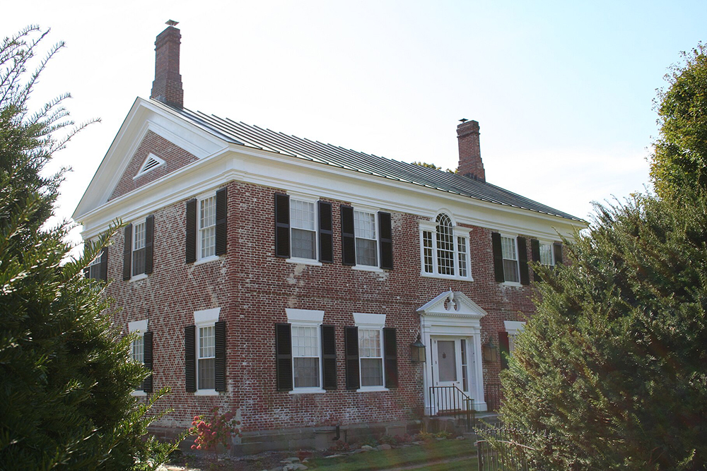 A historic two-story red brick colonial house with black shutters, white trim, and twin chimneys, surrounded by greenery under a clear sky