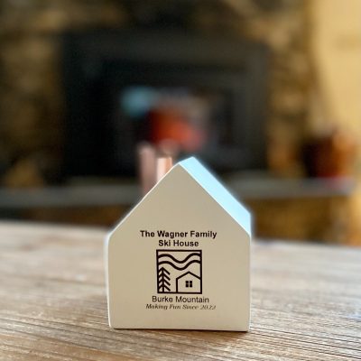 A little white wooden house candlestick holder with a copper chimney and a custom engraved decorative image and the words "The Wagner Family Ski House. Burke Mountain. Making Fun Since 2022."