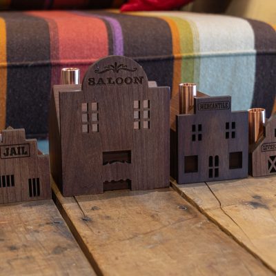 Four tiny wooden western village-themed candlestick holders in a row. A jail, saloon, mercantile, and livery stable.